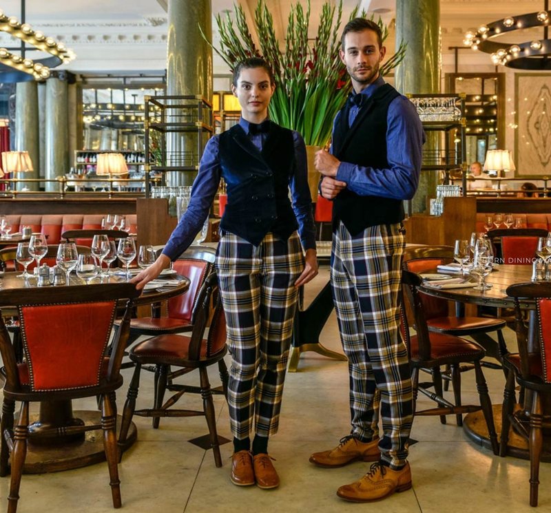 Two staff in checked trousers and waistcoats behind a bar