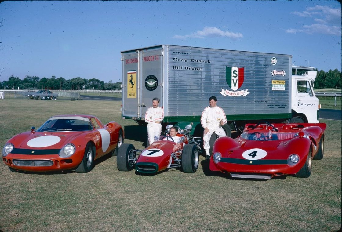 Three red Scuderia Veloce racing cars lined up on grass in front of the team transporter