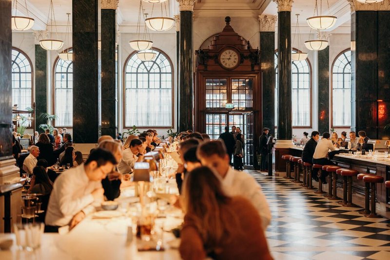 A grand hall of diners under arched windows at The Ned in London