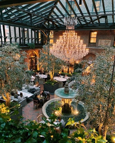A grand glass-roofed dining room with a chandelier, fountain and greenery